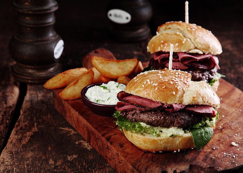 Gourmet Tasty Steak Burgers with Ham Slices on a Wooden Tray with Potato Wedges and Dipping Sauce.