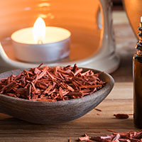 A bottle of sandalwood essential oil with sandalwood pieces and an aroma lamp in the background