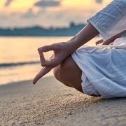 Woman in white clothes and a turban sits on a sandy beach at dawn and practices yoga